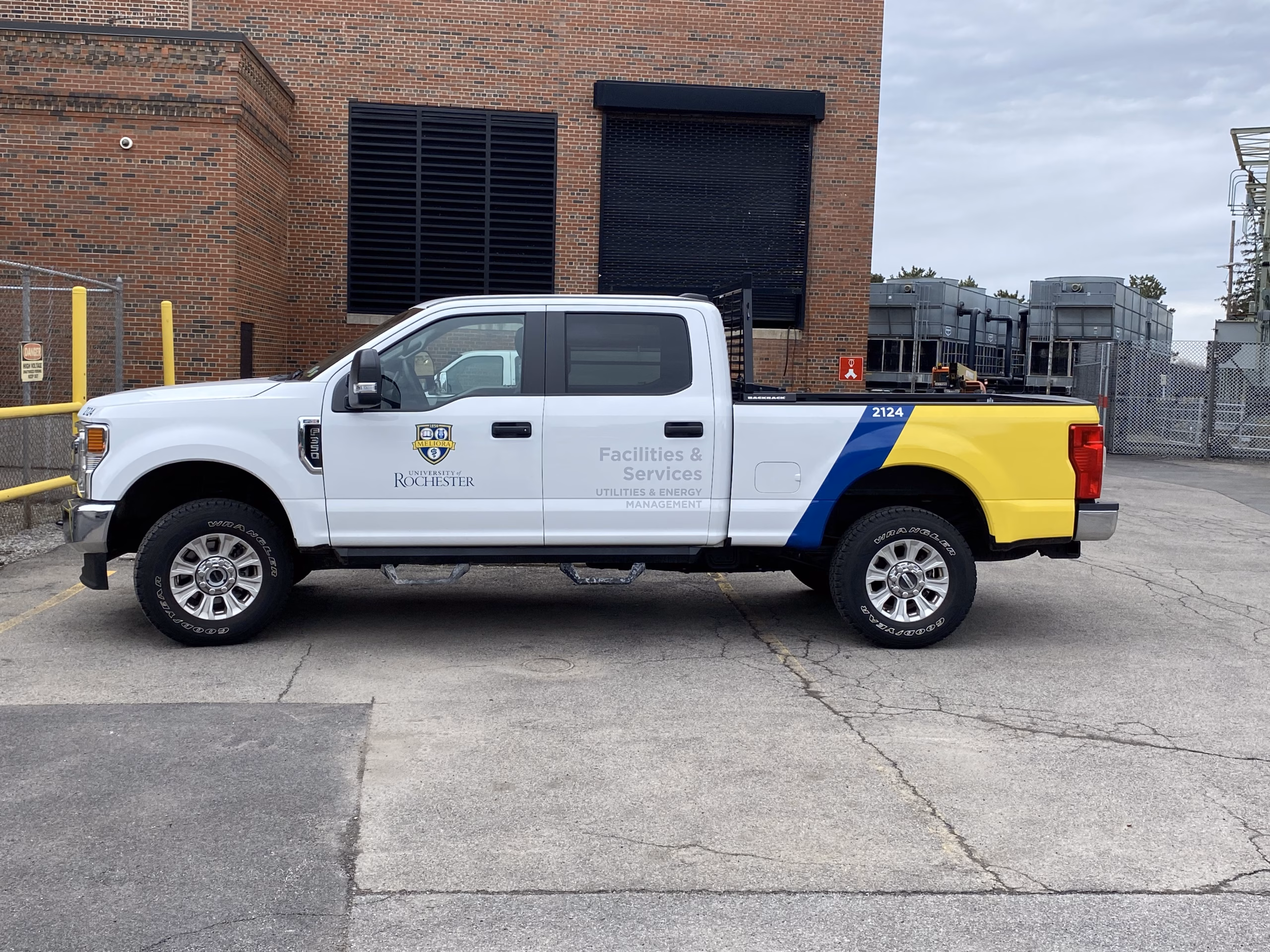 White, yellow and blue Facilities and Services truck parked in front of brick building.